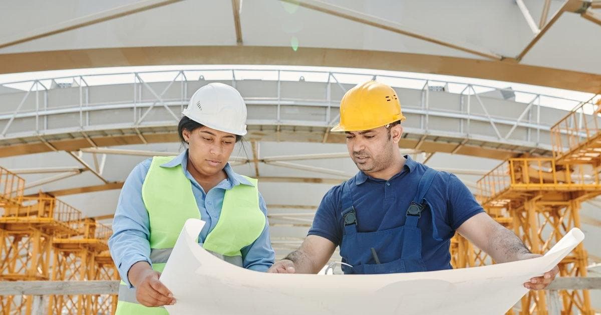 Dua ahli K3 konstruksi berhelm dan berompi keselamatan membaca gambar kerja di proyek gedung